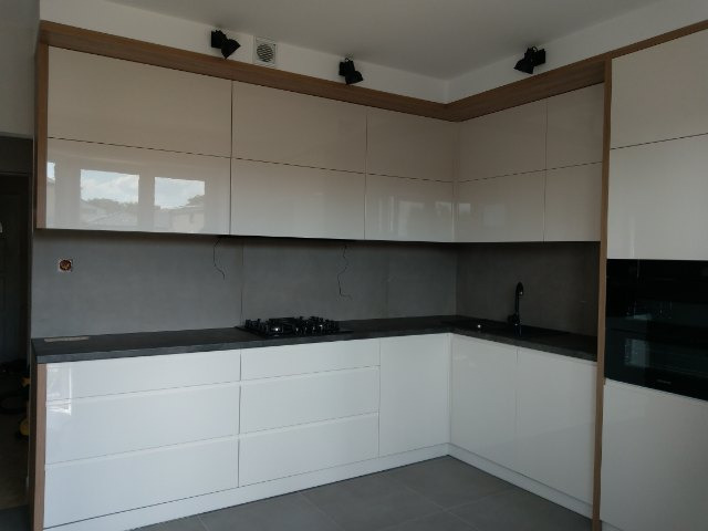 Nowoczesna, narożna kuchnia z białymi, lakierowanymi frontami, szarym blatem i podświetleniem LED, z widoczną płytą grzewczą i czarną armaturą.