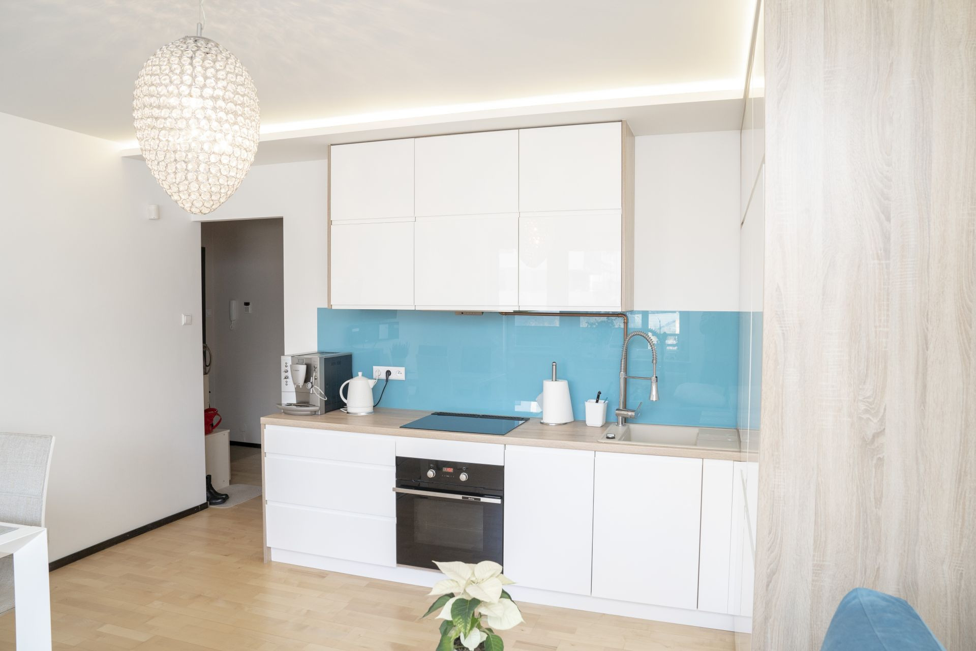 Nowoczesna, jasna kuchnia z białymi szafkami, turkusowym panelem szklanym, drewnianym blatem i ozdobnym żyrandolem w kształcie kuli.