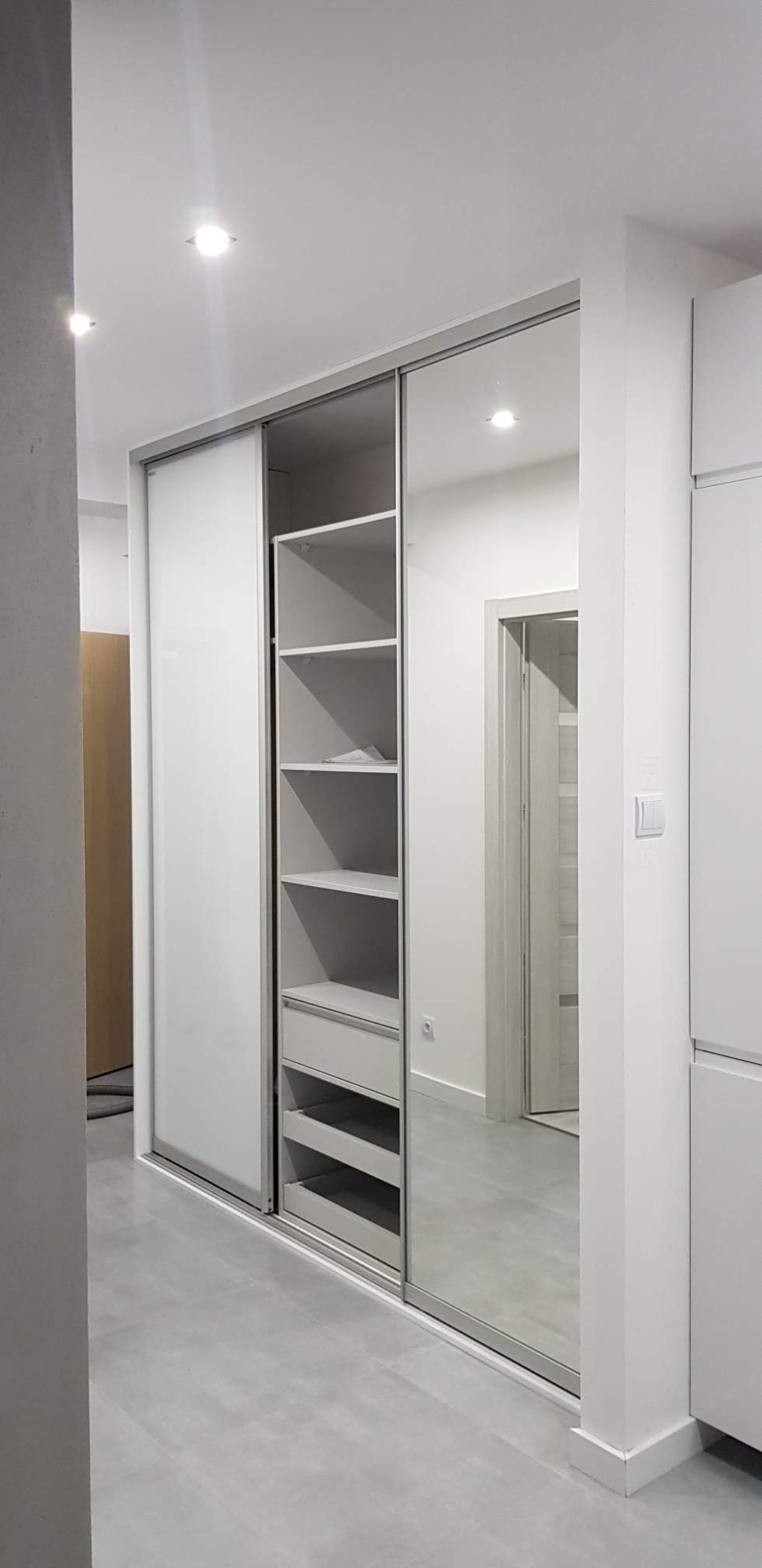 Wnętrze z zabudowaną szafą przesuwną z lustrzanymi drzwiami i otwartą częścią z półkami i szufladami w szaro-białej kolorystyce.