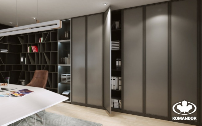 Szafa przesuwna z szarymi, matowymi frontami, częściowo otwarta, z widoczną zawartością półek: segregatory i książki, w tle biurko i regał z książkami, oświetlenie LED wewnątrz szafy, logo firmy...