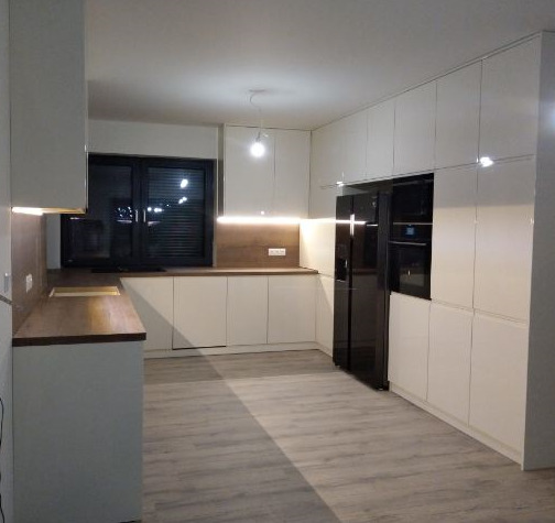 Nowoczesna kuchnia z białymi, lakierowanymi frontami, drewnianym blatem i czarną lodówką side-by-side, oświetlenie LED pod szafkami górnymi.