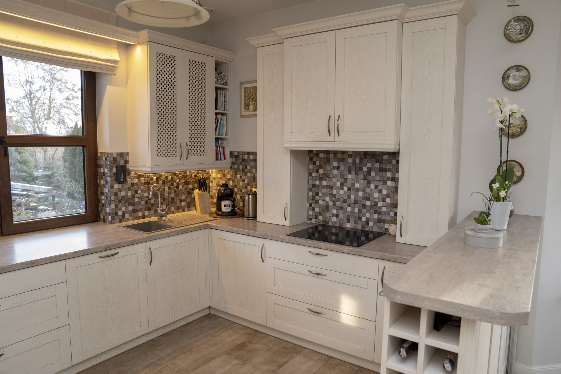 Jasna kuchnia z drewnianymi frontami, mozaiką nad blatem, oświetleniem LED pod szafkami i elementami dekoracyjnymi jak kwiaty i talerze na ścianie.