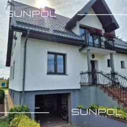 Biały, dwukondygnacyjny dom po termomodernizacji z ciemnymi oknami, dachem i elementami elewacji. Widoczne ocieplenie ścian, wymienione okna i nowa elewacja.