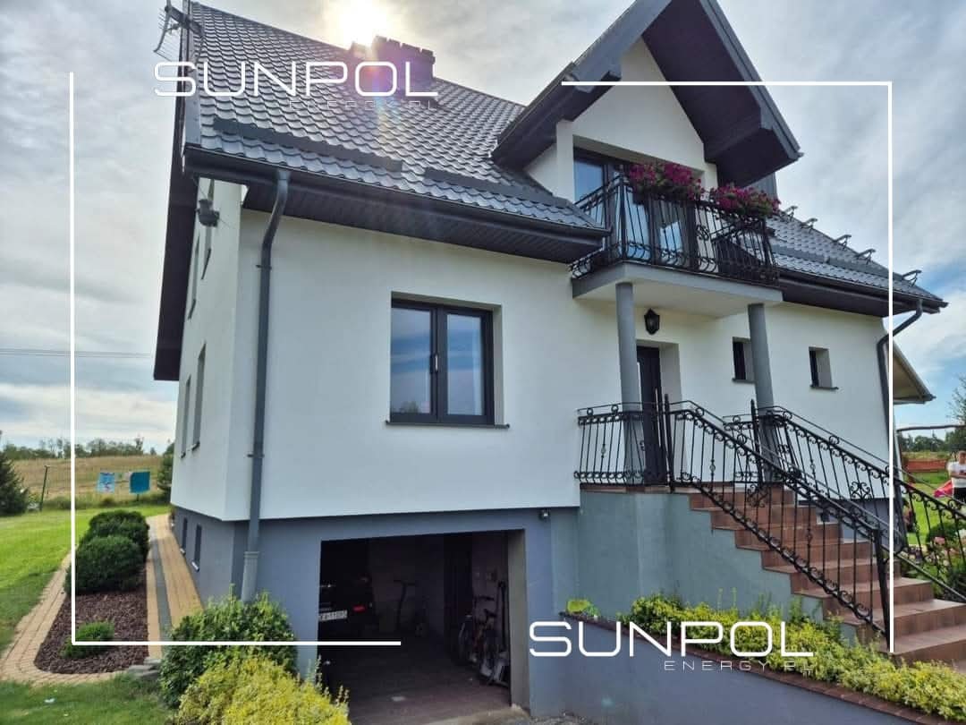 Biały, dwukondygnacyjny dom po termomodernizacji z ciemnymi oknami, dachem i elementami elewacji. Widoczne ocieplenie ścian, wymienione okna i nowa elewacja.