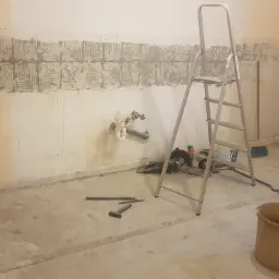 Pokój w trakcie remontu: ściana z częściowo usuniętymi płytkami, widoczne ślady po kleju, drabina, narzędzia budowlane i wiadro.