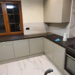 Nowoczesna kuchnia z szarymi szafkami, ciemnym blatem i marmurową podłogą po remoncie, widoczny odkurzacz sugerujący sprzątanie po pracach wykończeniowych.