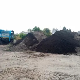 Dwie pryzmy ciemnej ziemi na placu budowy, z boku widoczna niebieska przesiewarka do ziemi i fragment traktora. W tle zabudowania mieszkalne.