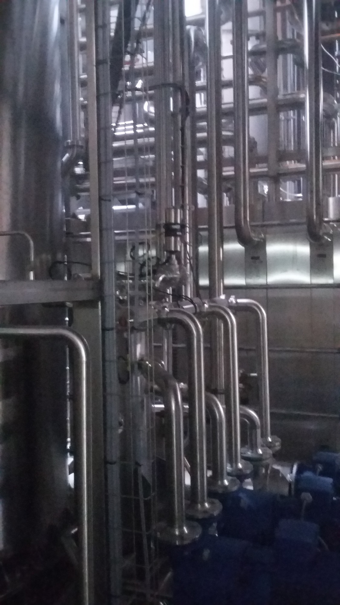 Kompleksowa instalacja rur ze stali nierdzewnej w zakładzie przemysłowym, widoczne liczne złącza i zawory, w tle zbiornik i oświetlenie przemysłowe.