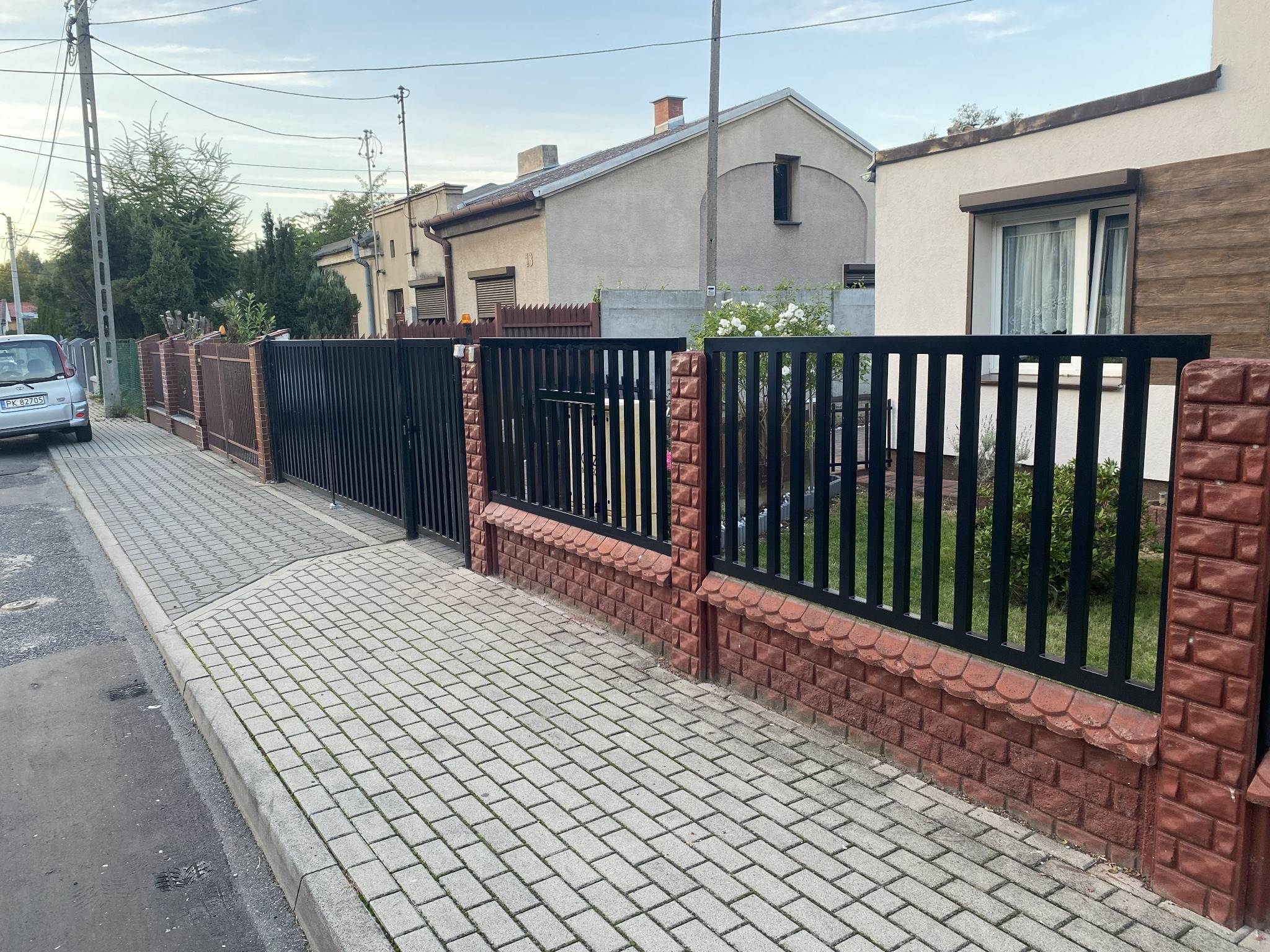 Czarne, metalowe ogrodzenie z furtką, osadzone na niskiej, ceglanej podmurówce. Widok z chodnika na posesję z zabudową jednorodzinną w tle.
