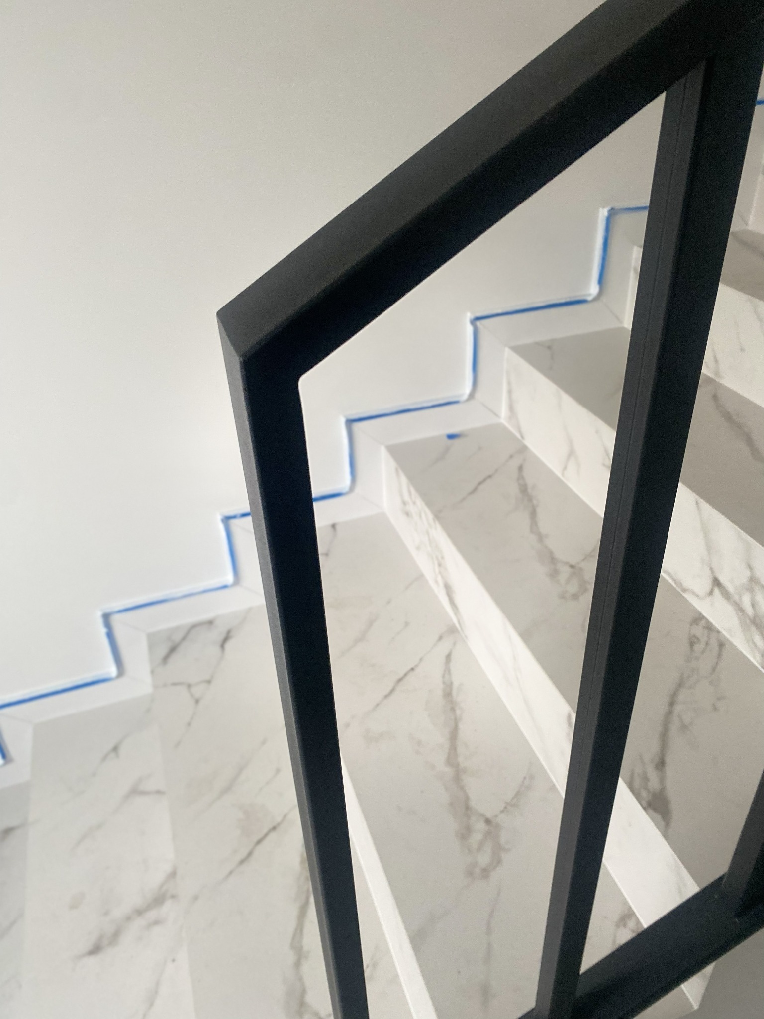 Nowoczesna balustrada schodowa z czarnych, metalowych profili na marmurowych schodach. Widok z góry, z niebieskim zabezpieczeniem malarskim przy ścianie.