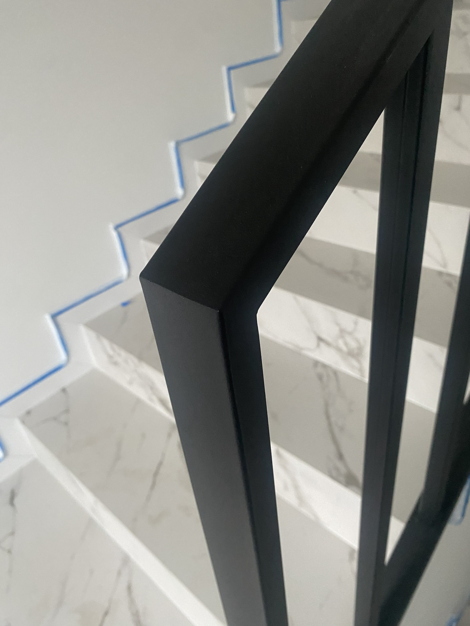Minimalistyczna, czarna balustrada schodowa z metalu, z bliska. Widoczne marmurowe stopnie zabezpieczone niebieską folią. Nowoczesny design.