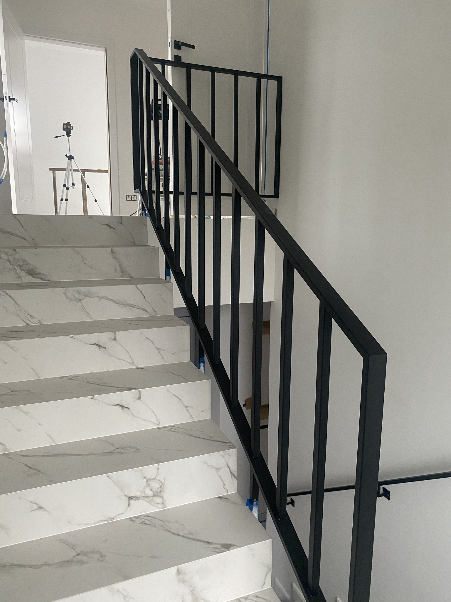 Nowoczesna, czarna balustrada schodowa z metalu, zamontowana na marmurowych schodach. W tle widoczny laser krzyżowy na statywie.