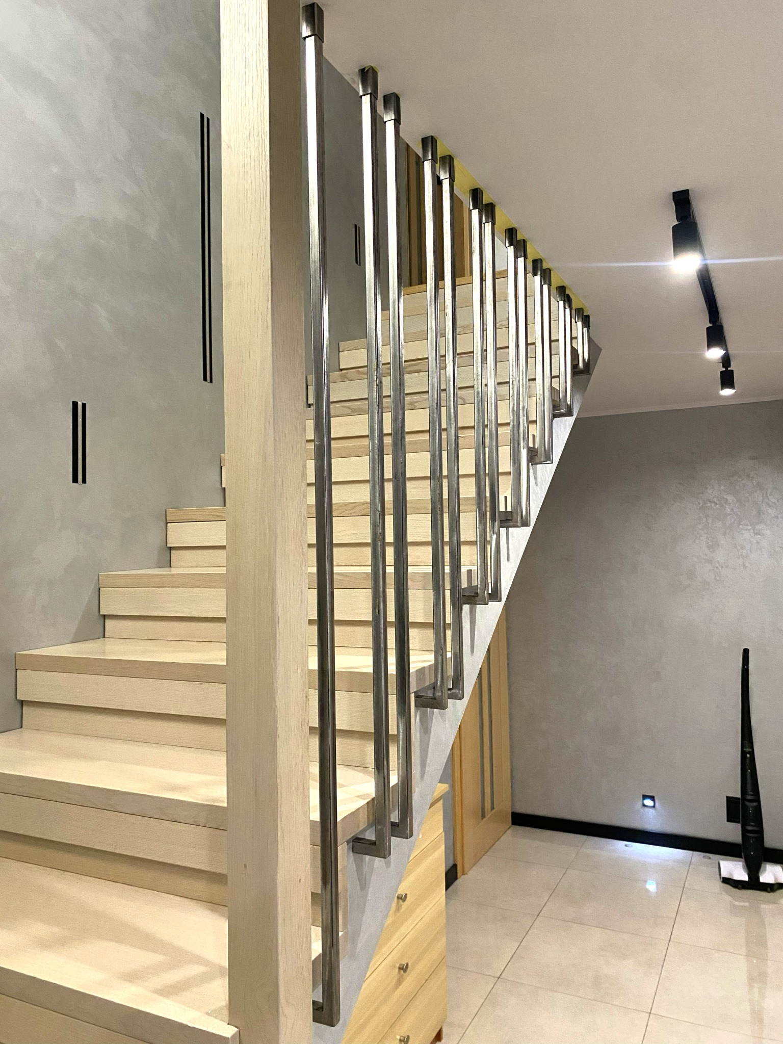 Nowoczesne schody z jasnego drewna i metalową balustradą, oświetlone punktowo, na tle szarej ściany z minimalistycznymi dekoracjami. Widok z dołu, podkreślający geometrię.