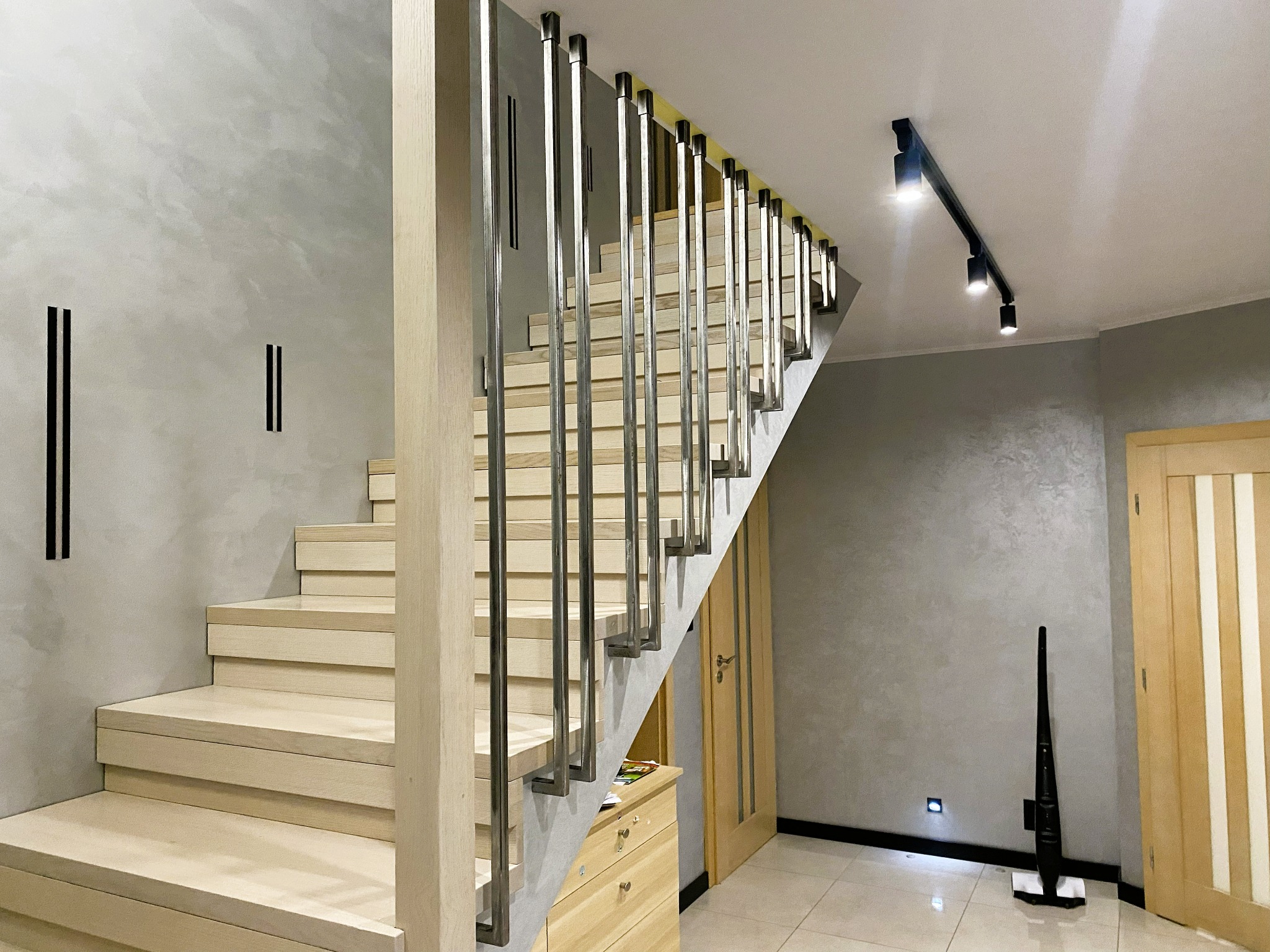 Nowoczesne schody z jasnego drewna z balustradą z pionowych, metalowych prętów. Minimalistyczny design, szara ściana z dekoracyjnymi elementami, oświetlenie punktowe.