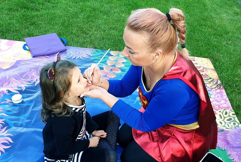 Kobieta w kostiumie superbohatera maluje twarz dziewczynce na kocu piknikowym na trawie, używając cienkiego pędzelka. Dziewczynka ma na głowie opaskę z uszami.