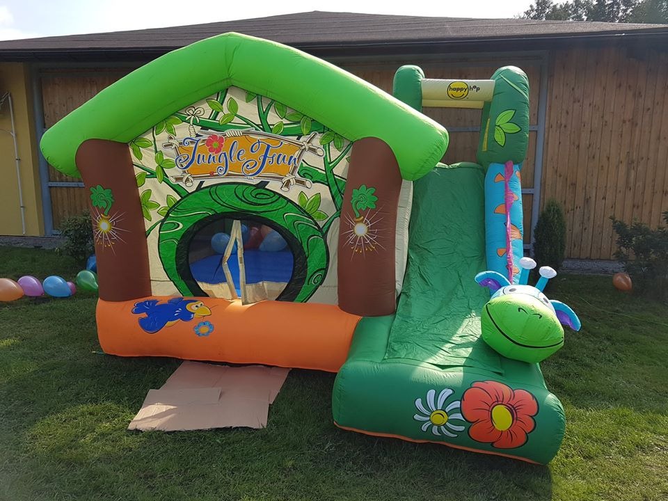 Kolorowy, dmuchany zamek ze zjeżdżalnią w motywie dżungli, z napisem 'Jungle Fun', ustawiony na trawie, z balonami w tle.