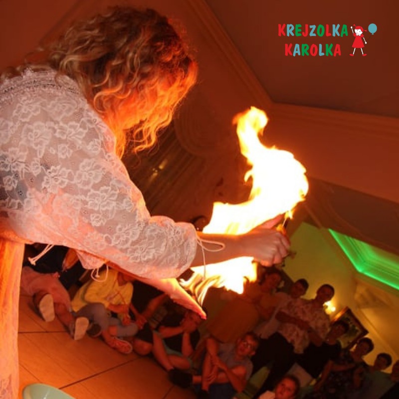 Osoba w koronkowej bluzce prezentuje płonący rekwizyt na tle siedzących dzieci i logo 'Krejzolka Karolka' w prawym górnym rogu.