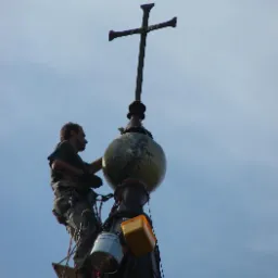 Pracownik na wysokościach, zabezpieczony linami, wykonuje prace konserwatorskie na iglicy kościoła zwieńczonej krzyżem; widoczny kubeł z farbą i pojemnik na narzędzia.