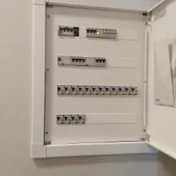 Otwarta, biała skrzynka elektryczna z zainstalowanymi bezpiecznikami i wyłącznikami różnicowoprądowymi, jeden z wyłączników świeci na zielono.