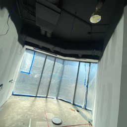 Music Gips - Przygotowanie pomieszczenia do prac wykończeniowych: ściany po szpachlowaniu, okna zabezpieczone folią, ślady na podłodze, sufit pomalowany na czarno, lampa zabezpieczona taśmą.