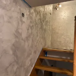 Ściana w korytarzu po nałożeniu dekoracyjnej, srebrzystej powłoki, widoczne schody drewniane i zabezpieczenia taśmą malarską.