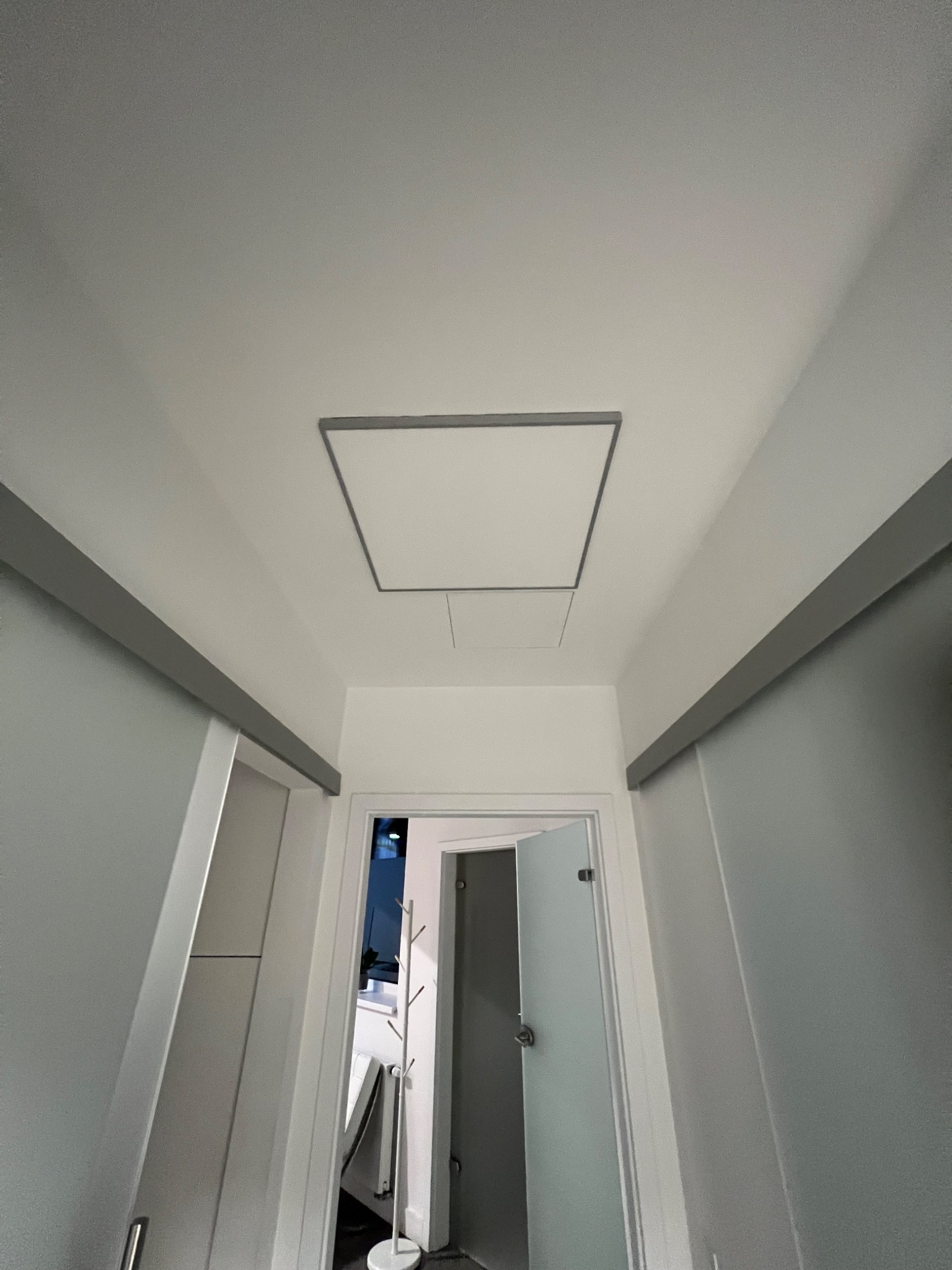 Biały sufit z zamontowanym kwadratowym włazem rewizyjnym, widok z dołu w kierunku otwartych drzwi z widocznym wieszakiem na ubrania.