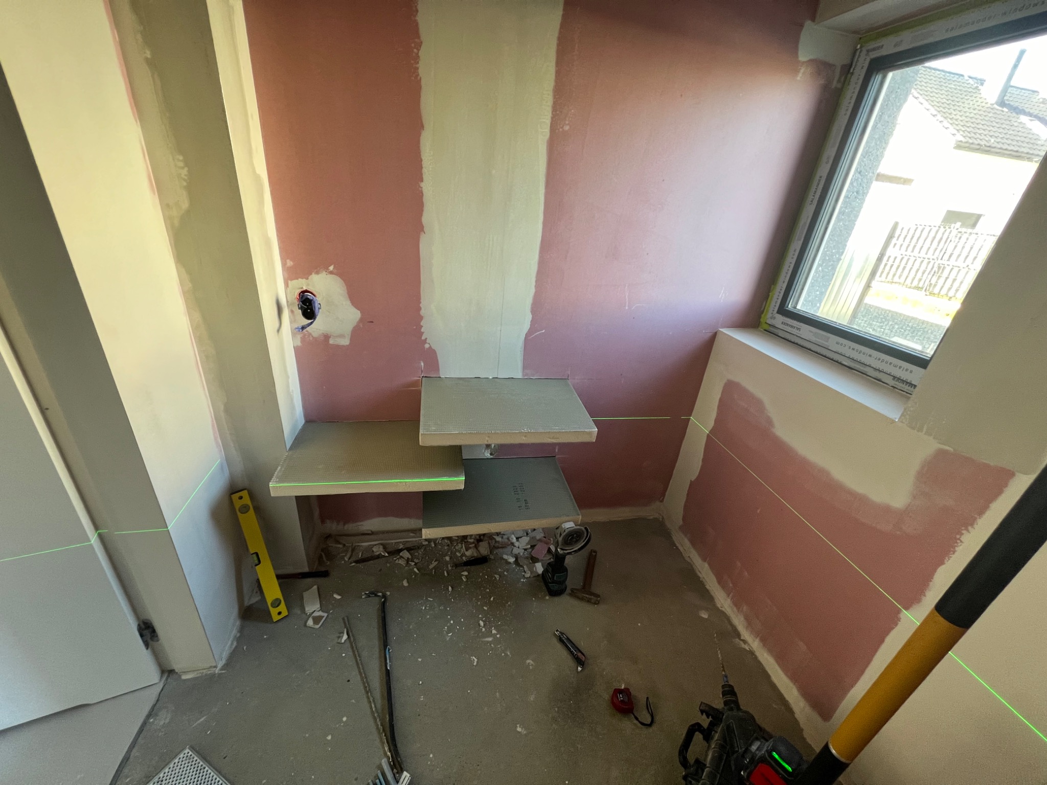 Przygotowanie ściany w Mucharzu do dalszych prac wykończeniowych, widoczne fragmenty nałożonej gładzi, surowe płyty na ścianie, poziomica laserowa wyznaczająca linie, narzędzia budowlane na podłodze.