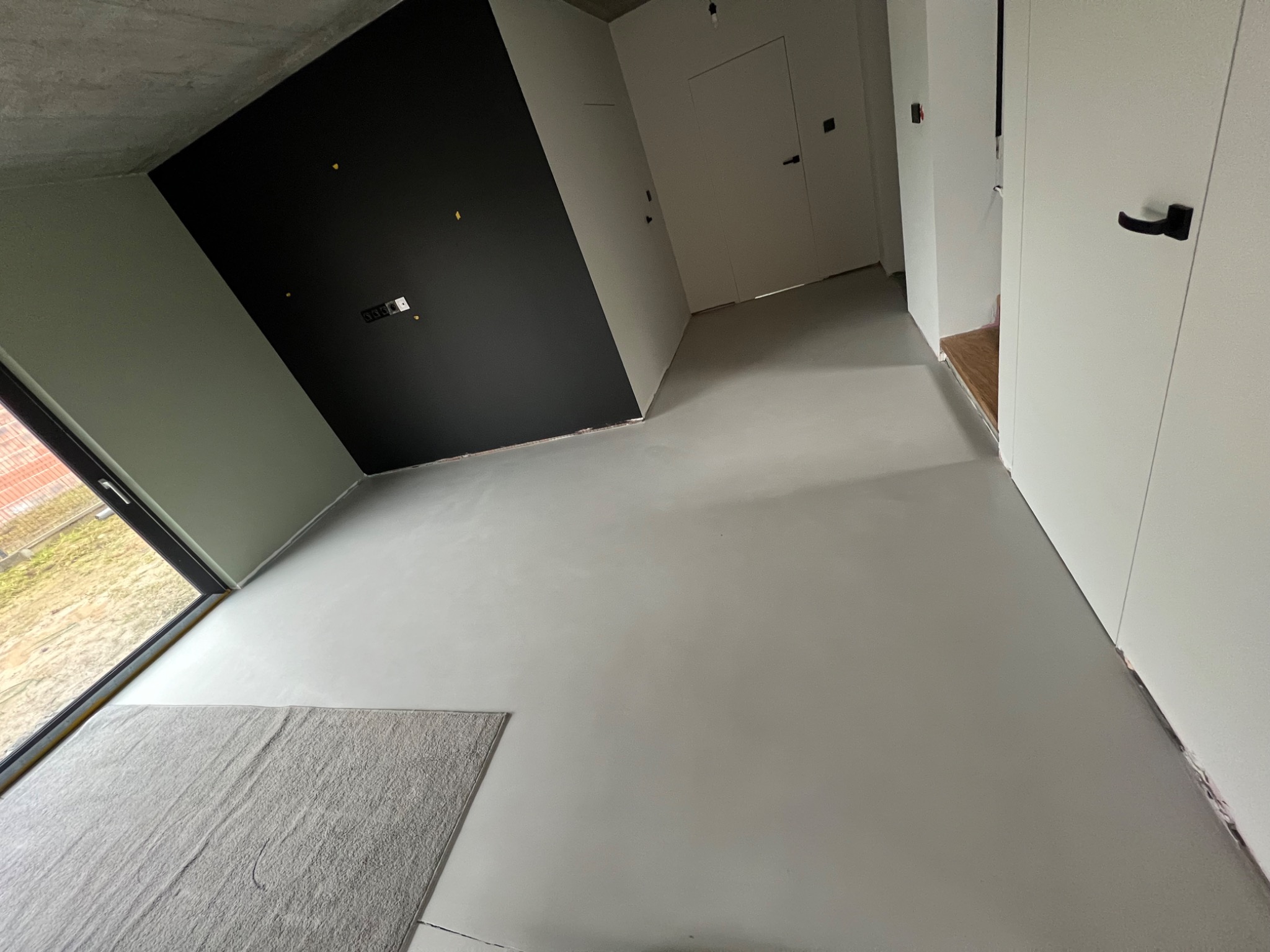 Nowoczesne wnętrze po remoncie: gładkie, szare ściany i podłoga, czarny akcent na ścianie, widoczne drzwi wewnętrzne i fragment okna z dywanem.