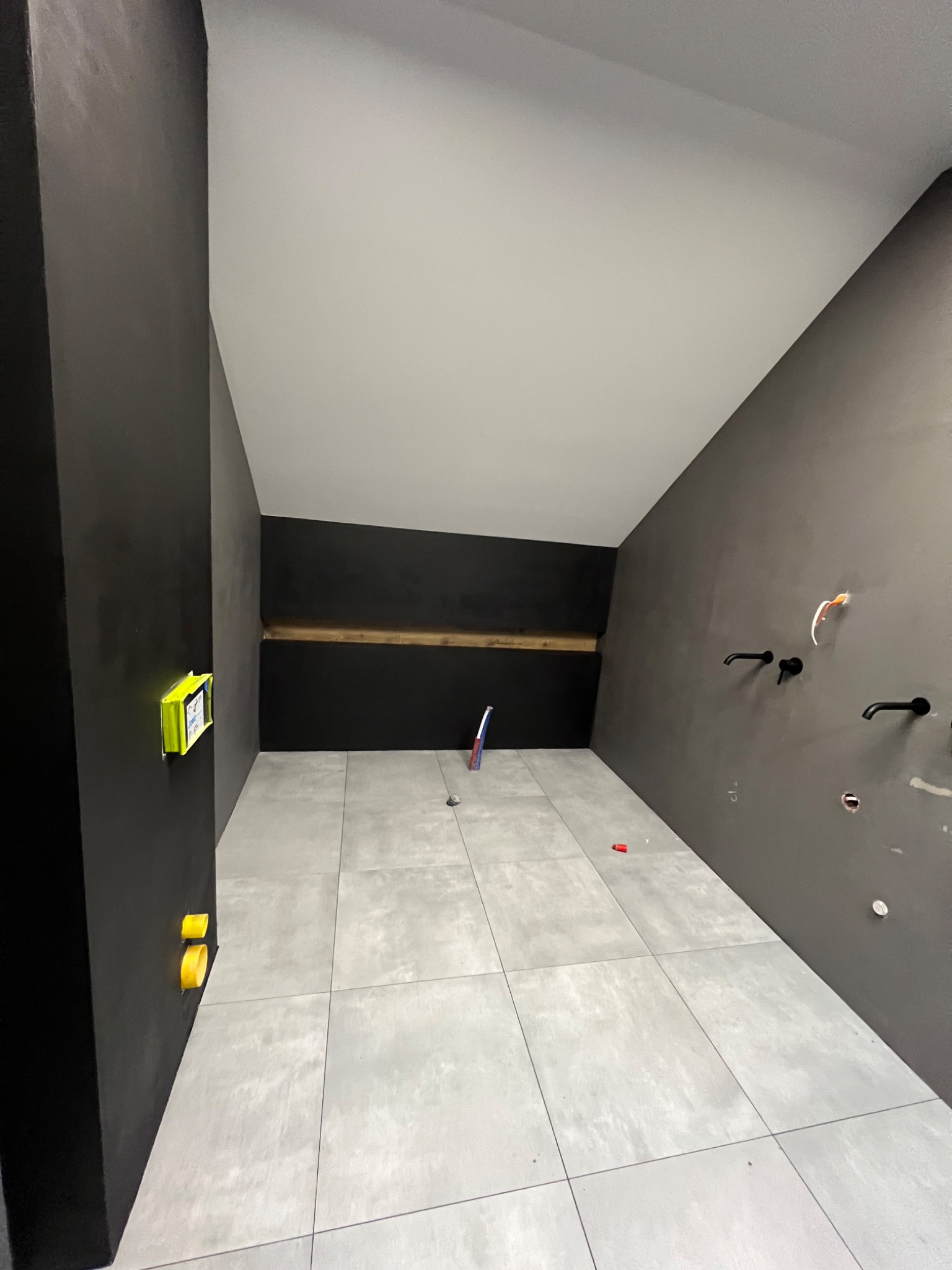 Wnętrze łazienki w trakcie wykańczania: szare ściany z widocznymi instalacjami wodno-kanalizacyjnymi, szare płytki podłogowe, poziomica na ścianie, częściowo pomalowany sufit.