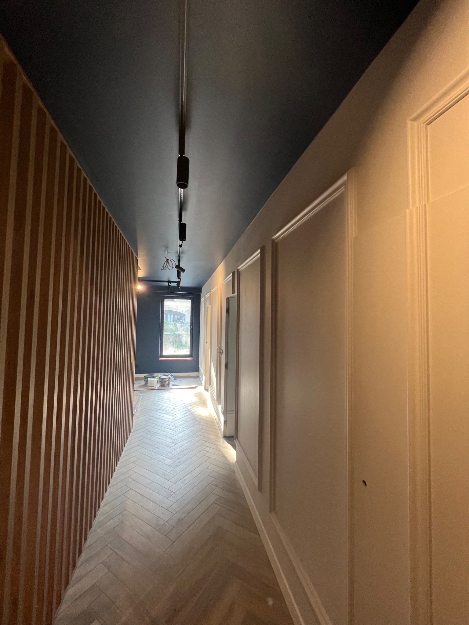 Korytarz po remoncie z ciemnoniebieskim sufitem, drewnianą ścianą z pionowych listew i jasną podłogą ułożoną w jodełkę. Na końcu korytarza widoczne okno i puszki z farbą.