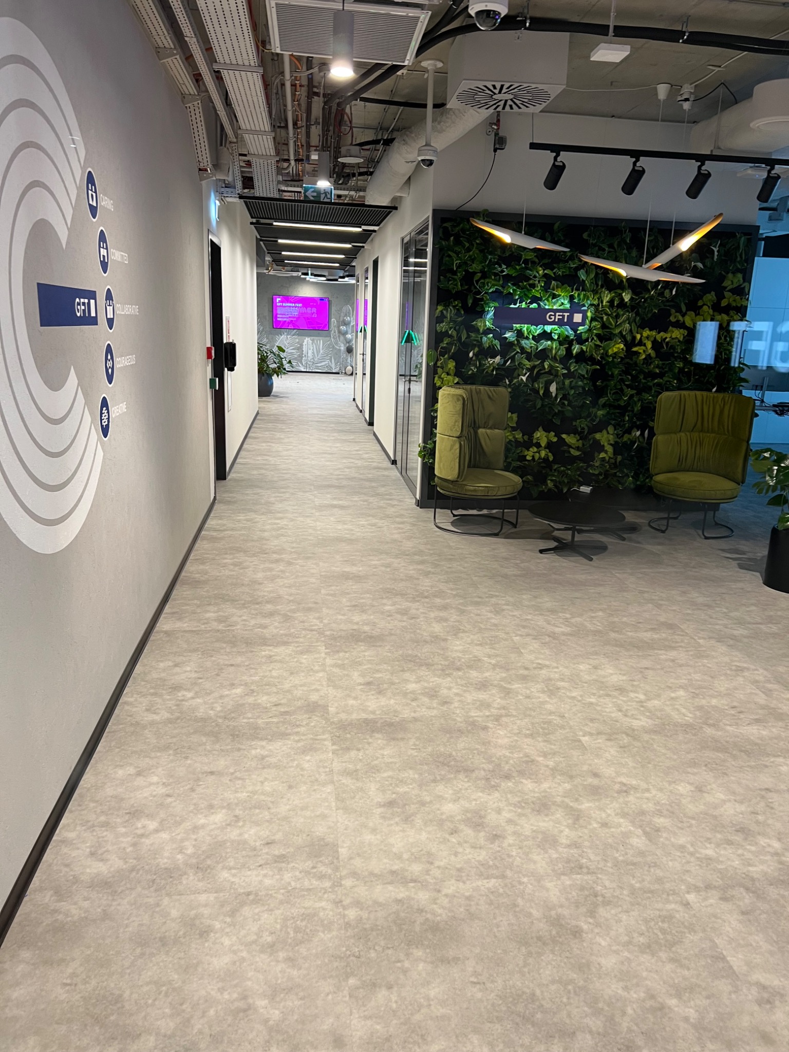 Nowoczesne wnętrze biurowe z długim korytarzem, szarą podłogą, ścianą z logo firmy i elementami graficznymi, oraz strefą relaksu z zieloną ścianą i fotelami w kolorze oliwkowym.