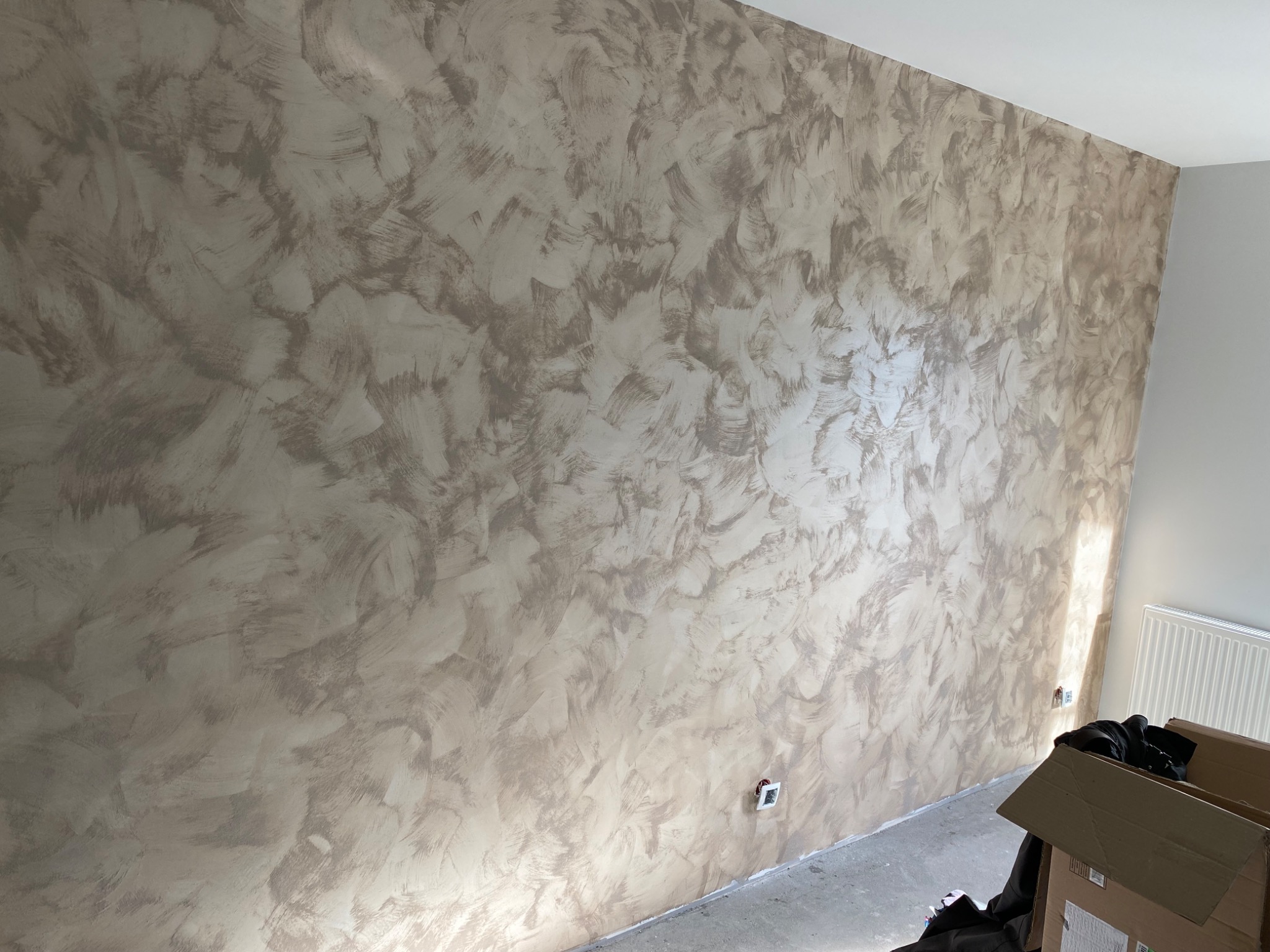 Ściana w odcieniach beżu i szarości z dekoracyjną fakturą imitującą pociągnięcia pędzla, widoczny fragment grzejnika i kartonowe pudło w rogu.