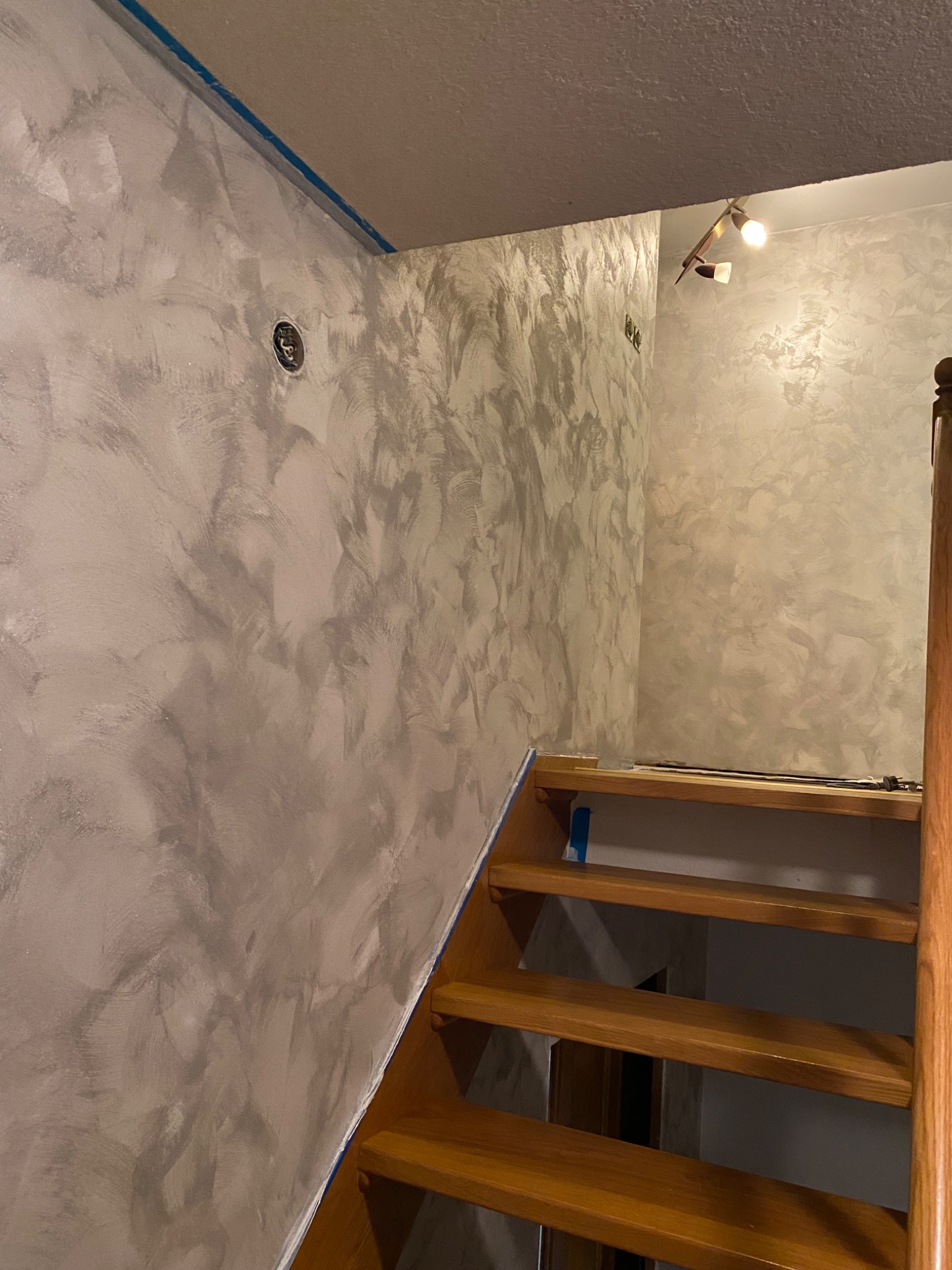 Ściana w korytarzu po nałożeniu dekoracyjnej, srebrzystej powłoki, widoczne schody drewniane i zabezpieczenia taśmą malarską.