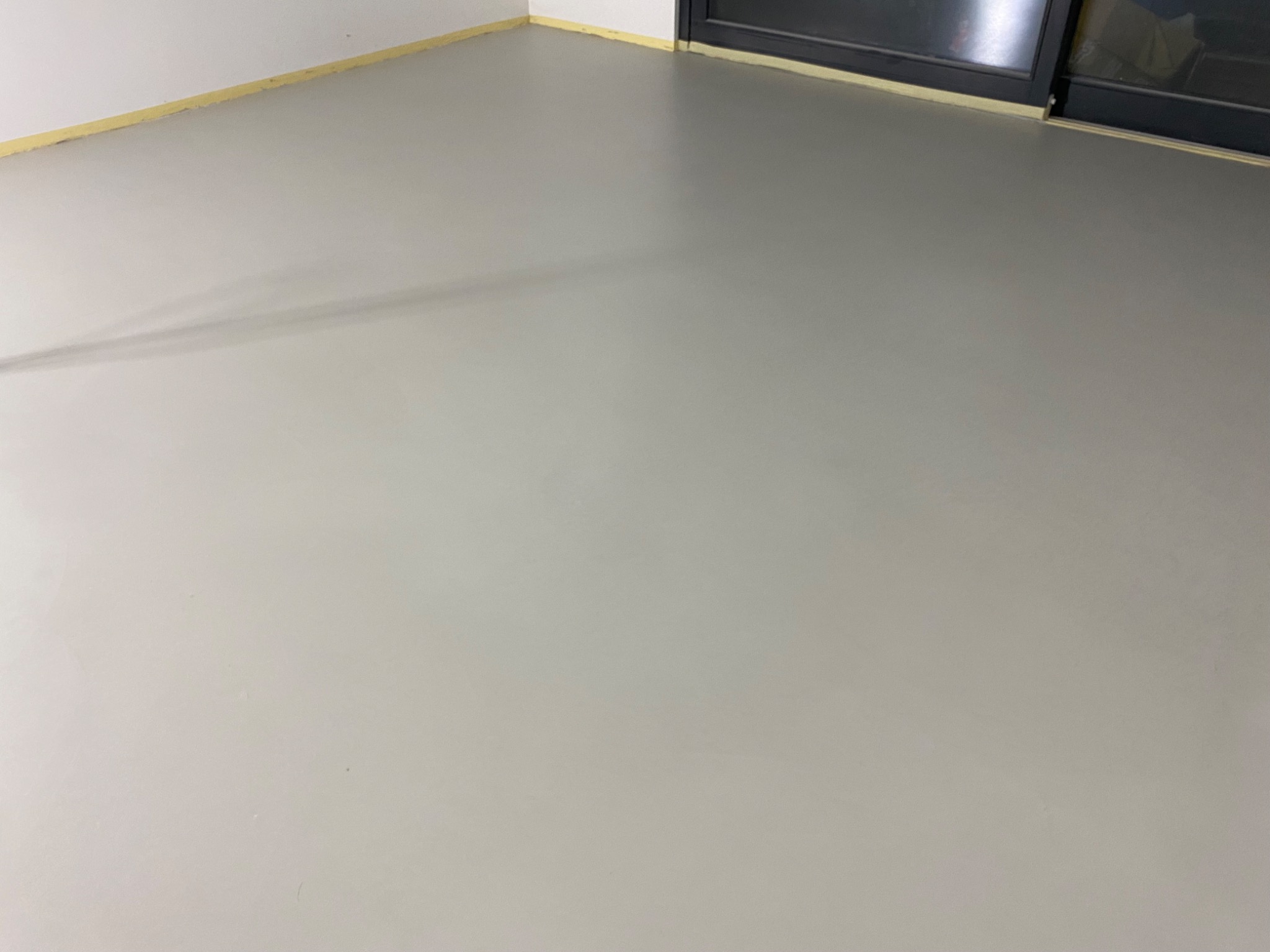 Świeżo wylana, gładka, jasnoszara posadzka w pomieszczeniu z oknem balkonowym i taśmą malarską zabezpieczającą ścianę przy podłodze.