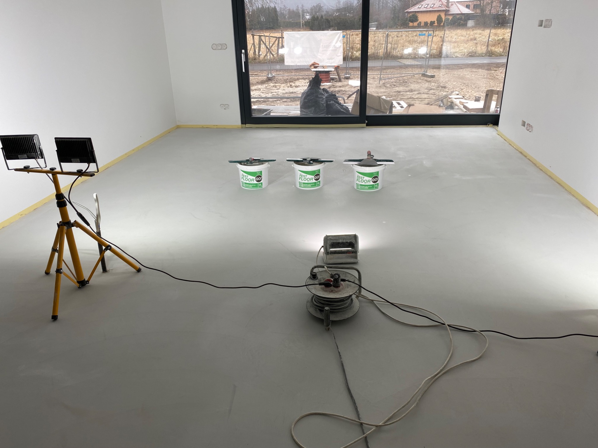 Świeżo wylana, jasnoszara posadzka samopoziomująca w nowym budynku, oświetlona lampami budowlanymi, z trzema wiadrami z mieszanką na posadzki i narzędziami do rozprowadzania w tle, zabezpieczone...