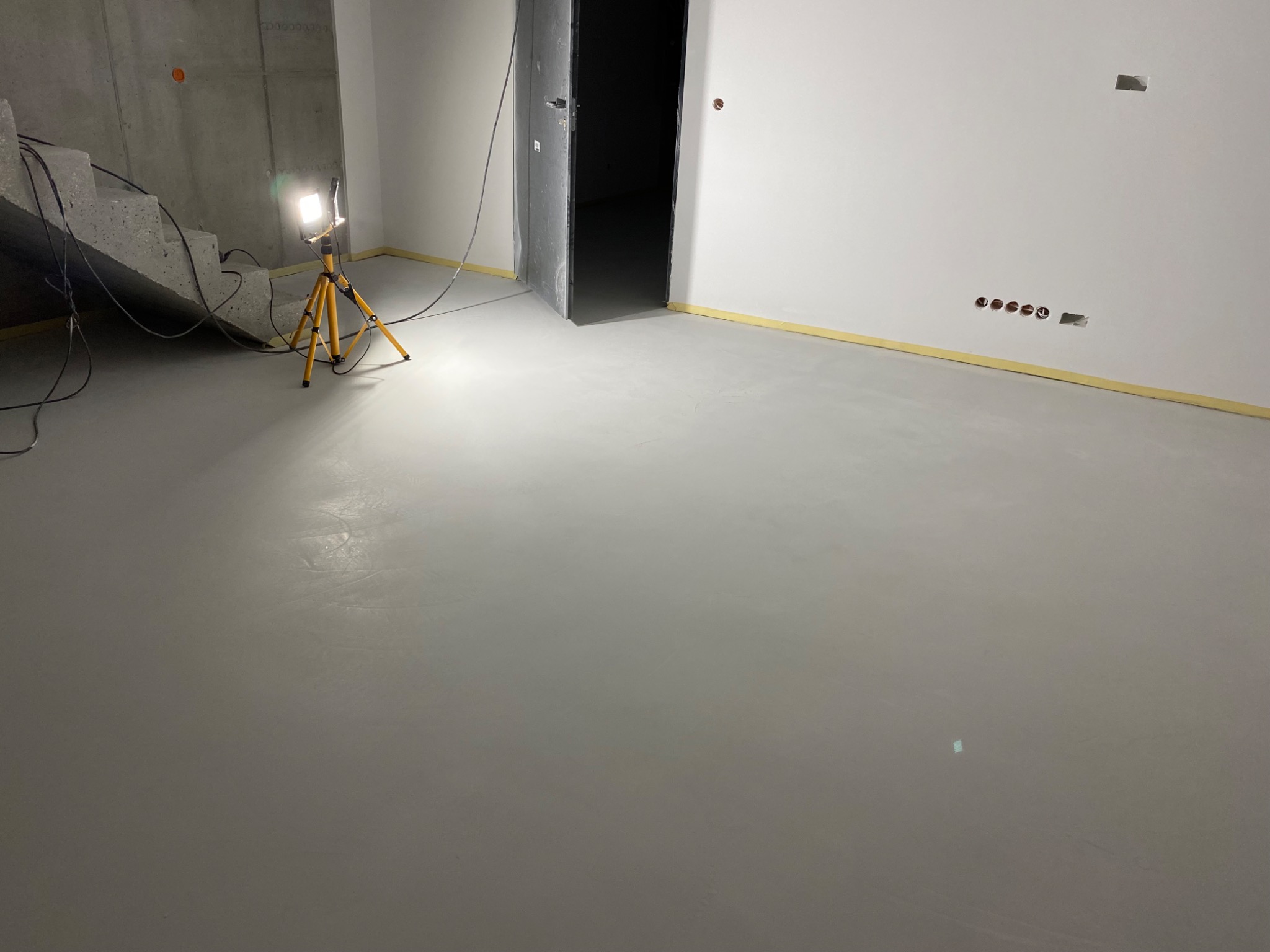 Wypoziomowana podłoga w surowym wnętrzu, oświetlona lampą na statywie, z widocznymi schodami w tle i taśmą malarską przy ścianach.