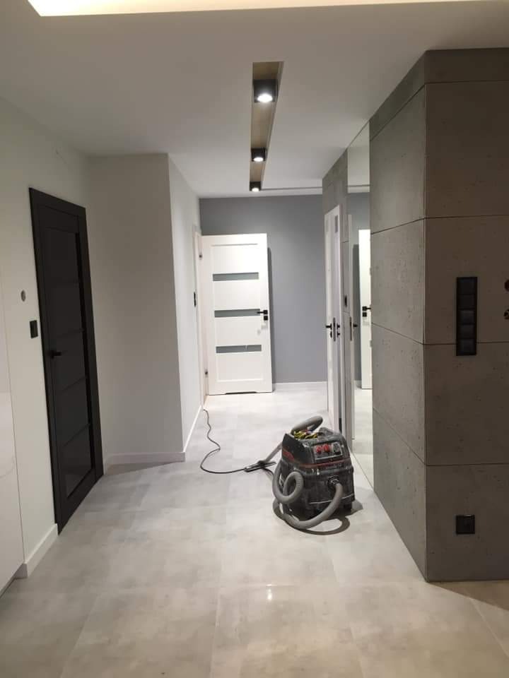 Korytarz po remoncie z widocznym odkurzaczem przemysłowym, szare płytki podłogowe, czarne drzwi i oświetlenie sufitowe punktowe.