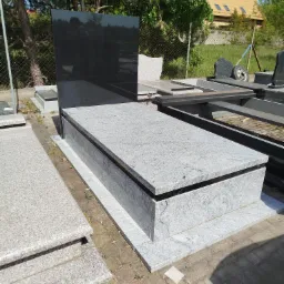 Nowy, szary nagrobek z czarną tablicą, ustawiony na placu z kostki brukowej, w tle inne nagrobki i zieleń.