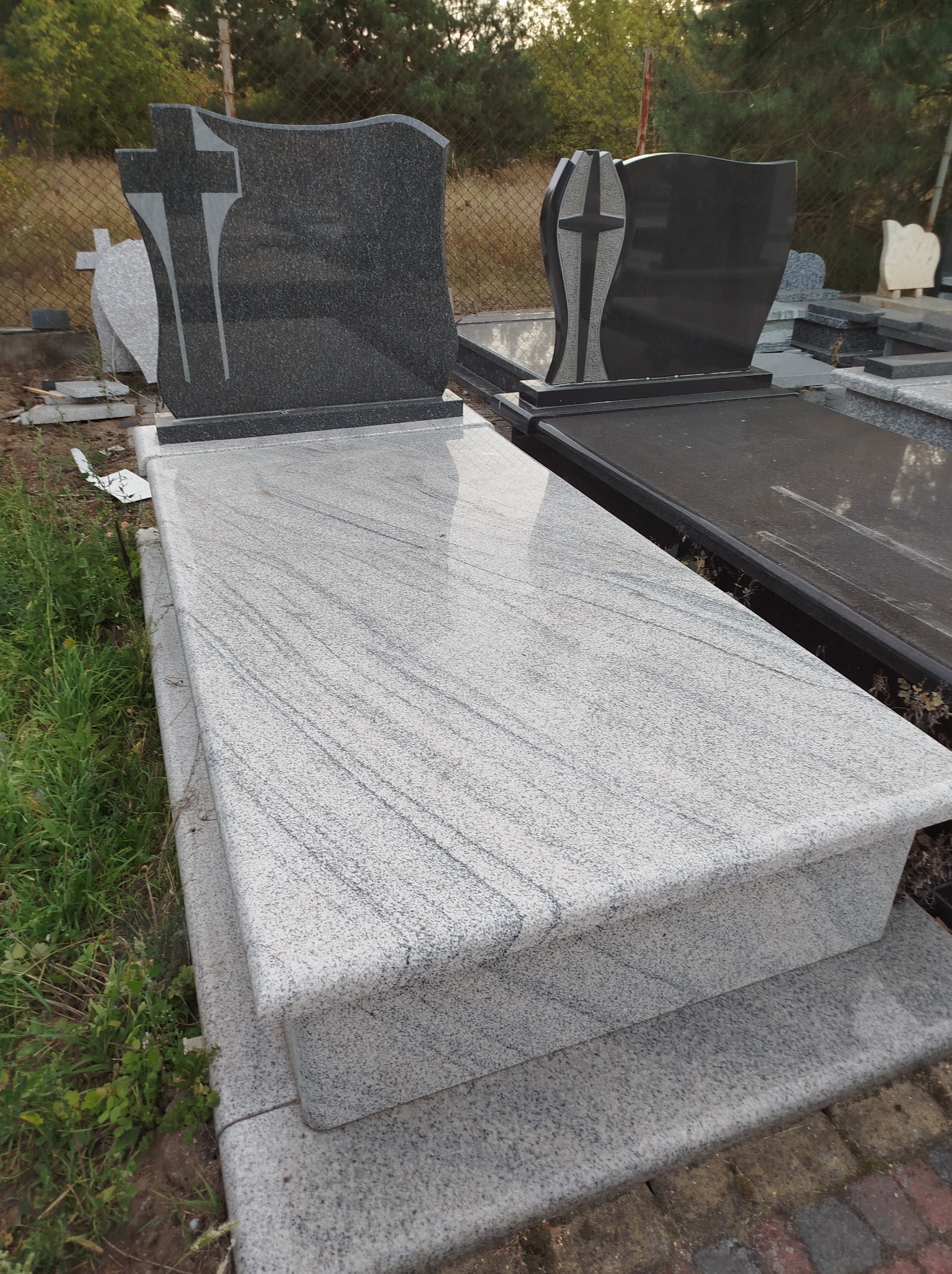 Jasnoszary, polerowany nagrobek z ciemnoszarą płytą nagrobną z motywem krzyża, widoczny na ekspozycji wśród innych nagrobków.