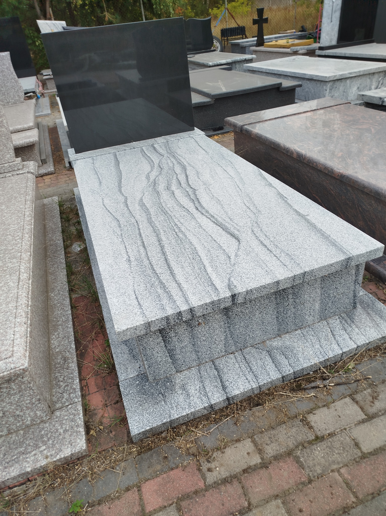 Nowy, jasnoszary nagrobek z czarną płytą, eksponowany na zewnątrz, z widocznymi wzorami w kamieniu i otoczeniem innych nagrobków oraz kostki brukowej.