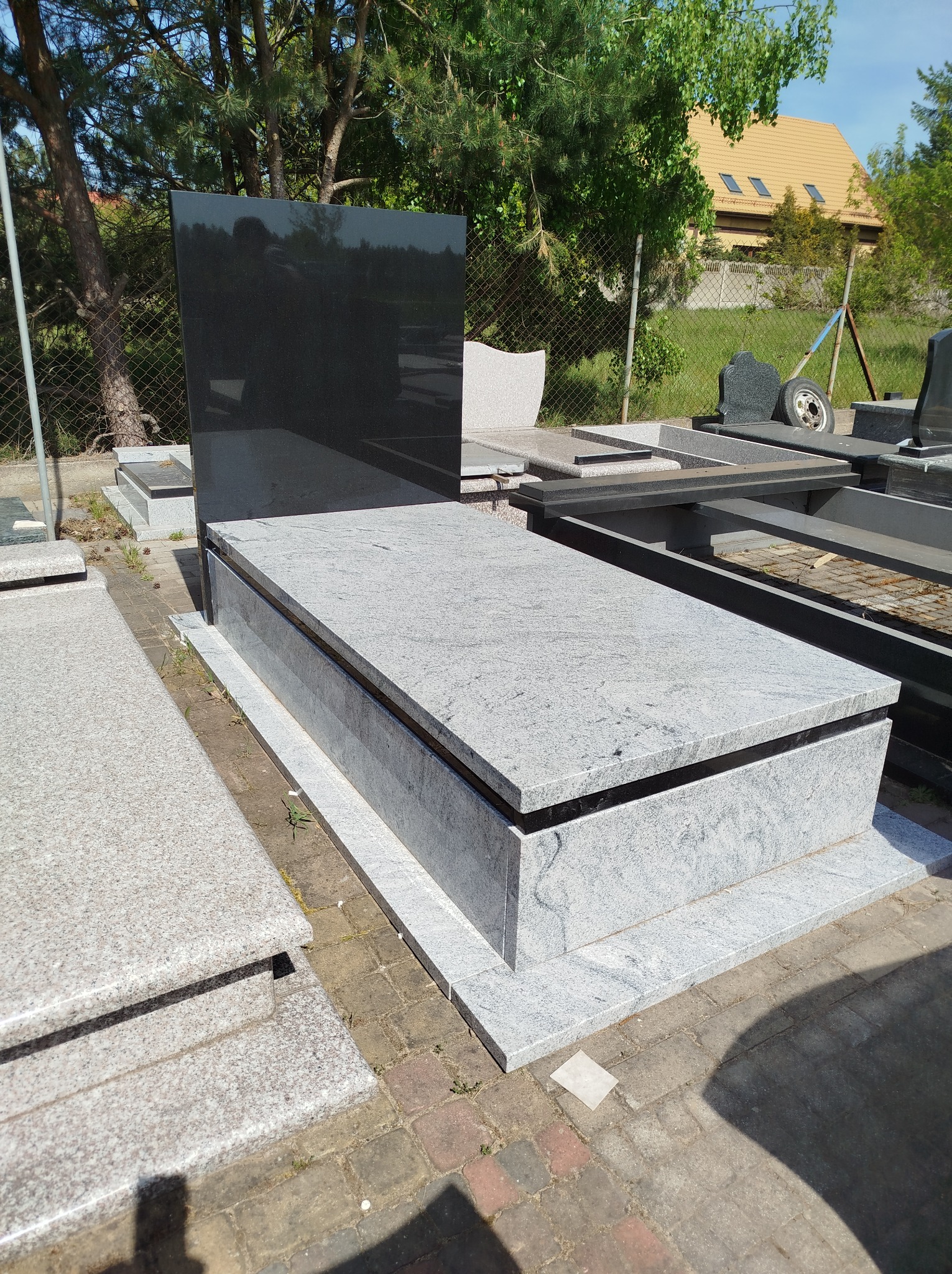 Nowy, szary nagrobek z czarną tablicą, ustawiony na placu z kostki brukowej, w tle inne nagrobki i zieleń.