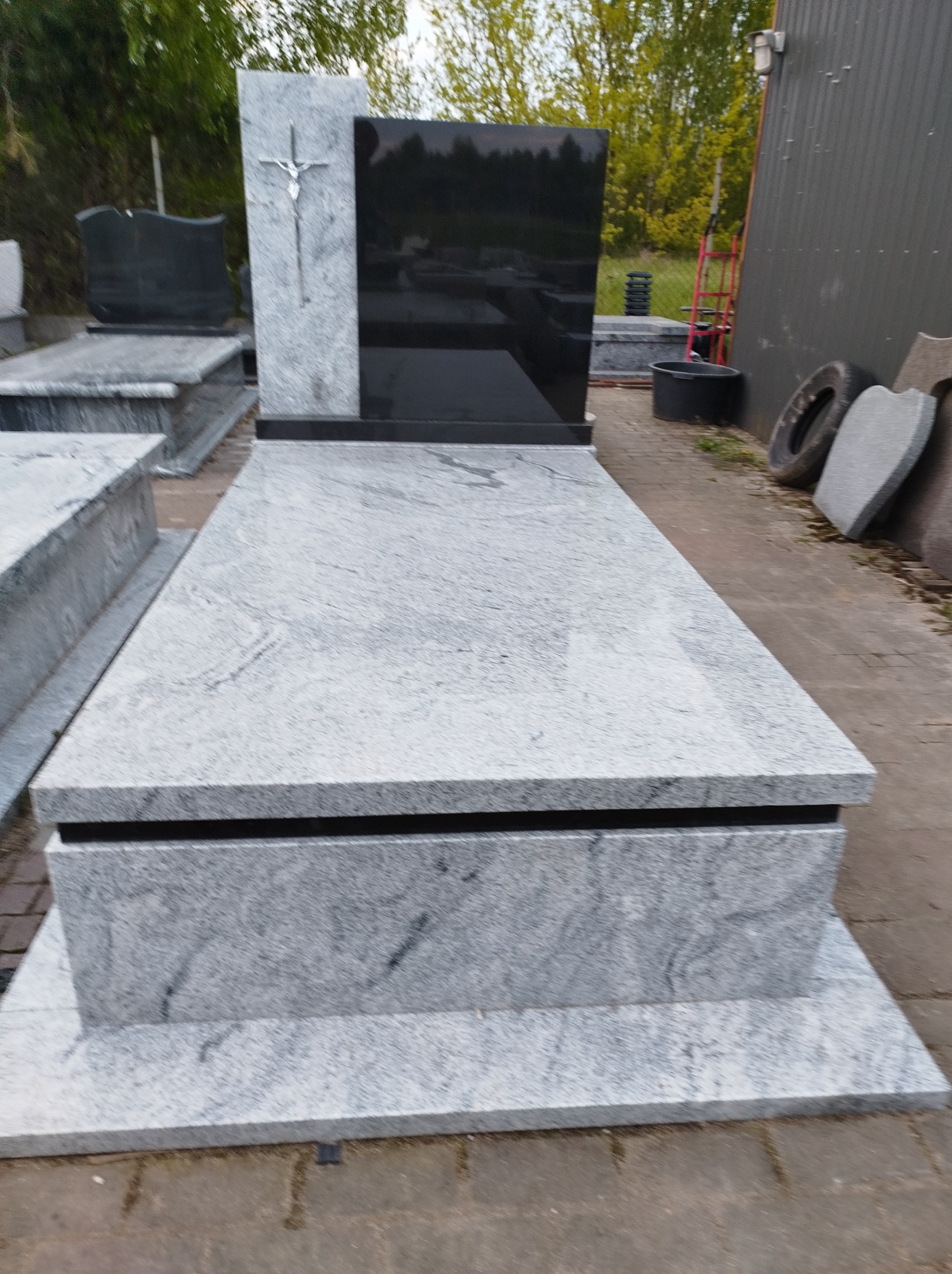 Jasnoszary, granitowy pomnik z czarną płytą nagrobną i srebrnym krzyżem, ustawiony na placu z kostki brukowej, widoczne inne nagrobki w tle.