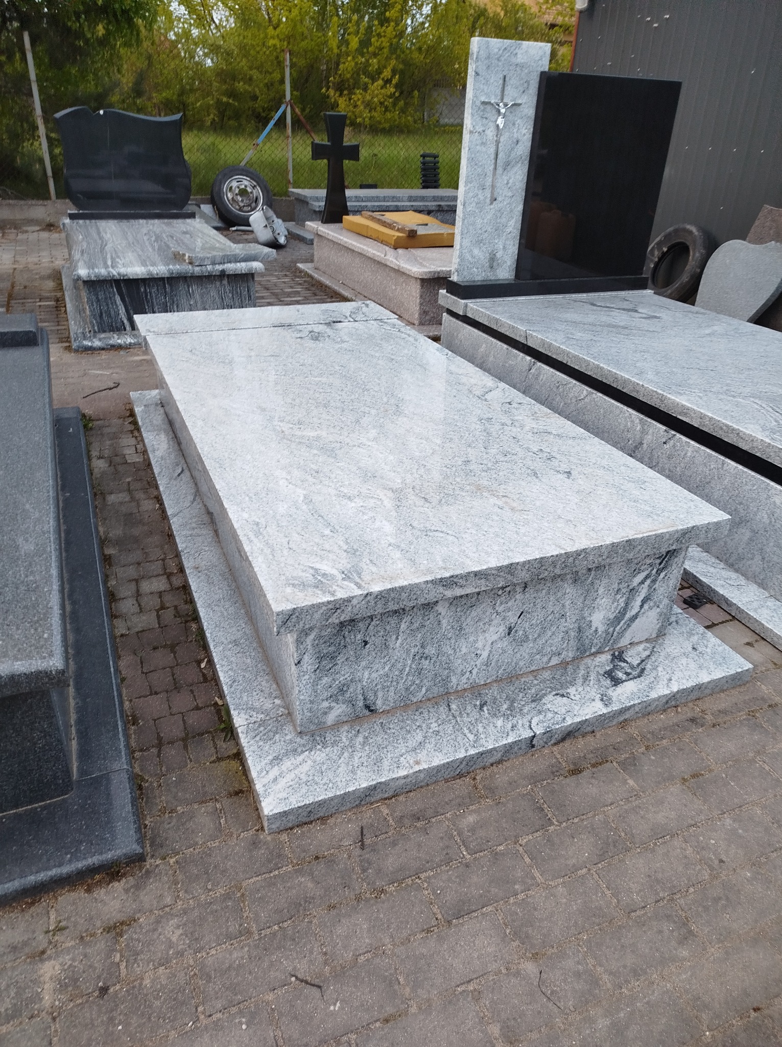 Ekspozycja nagrobków z jasnego granitu o różnej stylistyce, na zewnątrz, z oponą samochodową w tle.