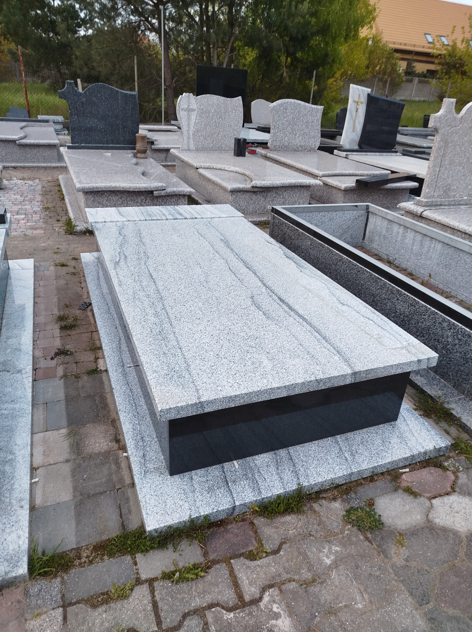 Wystawa nagrobków z jasnego granitu z czarną podstawą, widoczne inne pomniki w tle, ułożone na kostce brukowej.