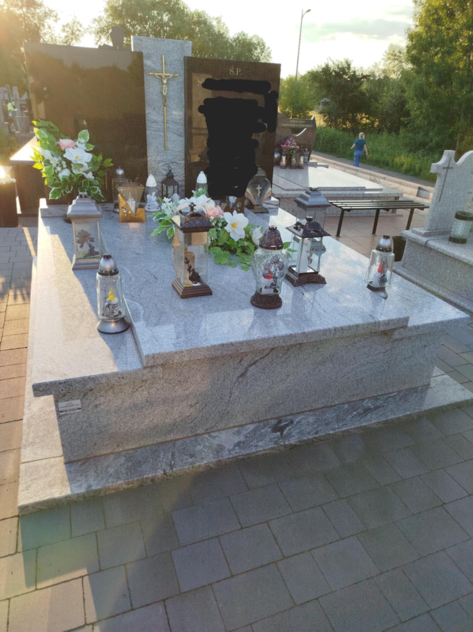Granitowy nagrobek z kwiatami i lampionami na cmentarzu w słoneczny dzień, widok z boku, w tle osoba oddalająca się alejką.