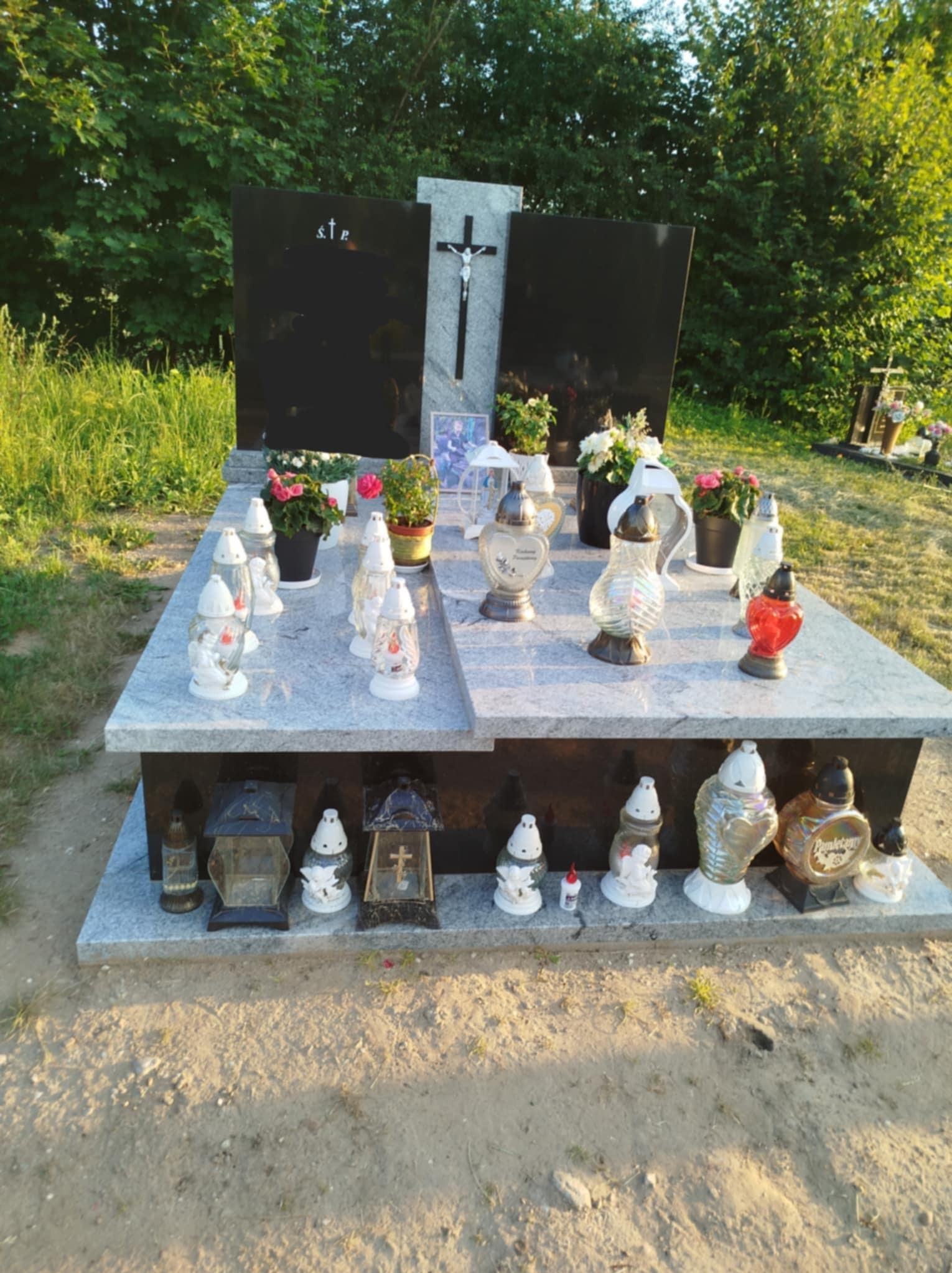 Granitowy pomnik z czarnymi tablicami, ozdobiony kwiatami w doniczkach i różnorodnymi lampionami, ustawiony na cmentarzu w słoneczny dzień.