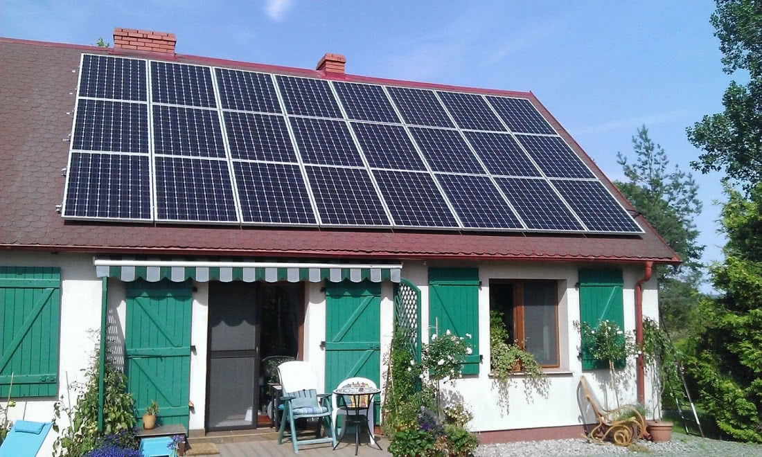 Instalacja fotowoltaiczna na dachu domu jednorodzinnego z czerwoną dachówką, zielonymi okiennicami i markizą nad tarasem.