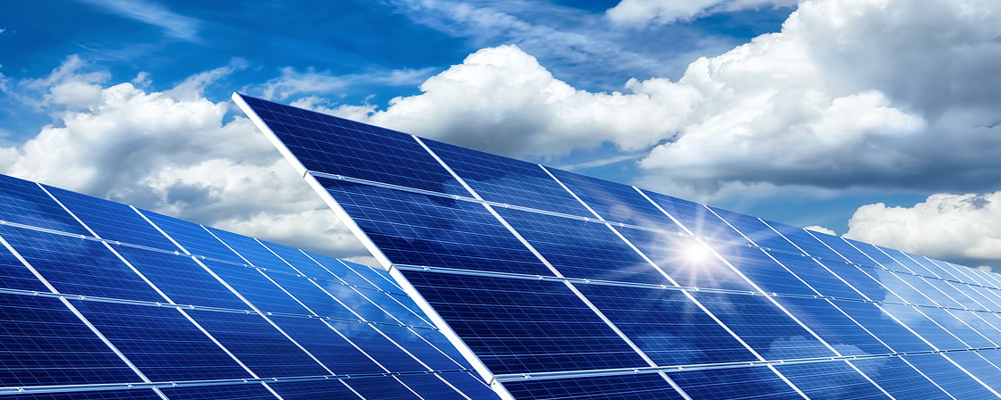 Błękitne panele słoneczne odbijają światło słoneczne na tle błękitnego nieba z białymi chmurami.
