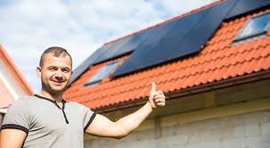 Uśmiechnięty mężczyzna kciukiem w górę na tle dachu z panelami słonecznymi i oknami dachowymi w słoneczny dzień.