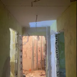 Korytarz w trakcie remontu: widoczne ściany z resztkami tapet, sufit z zamontowaną płytą karton gips, odsłonięte instalacje elektryczne i hydrauliczne, gruz na podłodze.